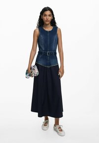 Denim sleeveless top with zipper, belted waist, and frayed hem combined with a long navy skirt. Model holds a patterned clutch.