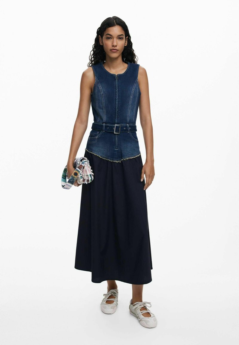 Denim sleeveless top with zipper, belted waist, and frayed hem combined with a long navy skirt. Model holds a patterned clutch.