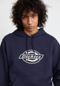 Haut à capuche bleu marine avec cordon de serrage. Présente un grand logo blanc brodé avec un motif en corde. Le tissu semble doux et texturé.