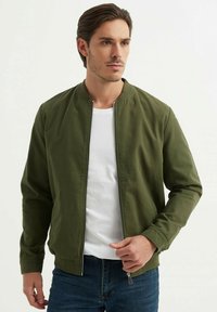 Veste bomber verte olive avec un col rond et fermeture éclair, associée à un t-shirt blanc et un jean bleu. Tissu en coton doux, design minimaliste.