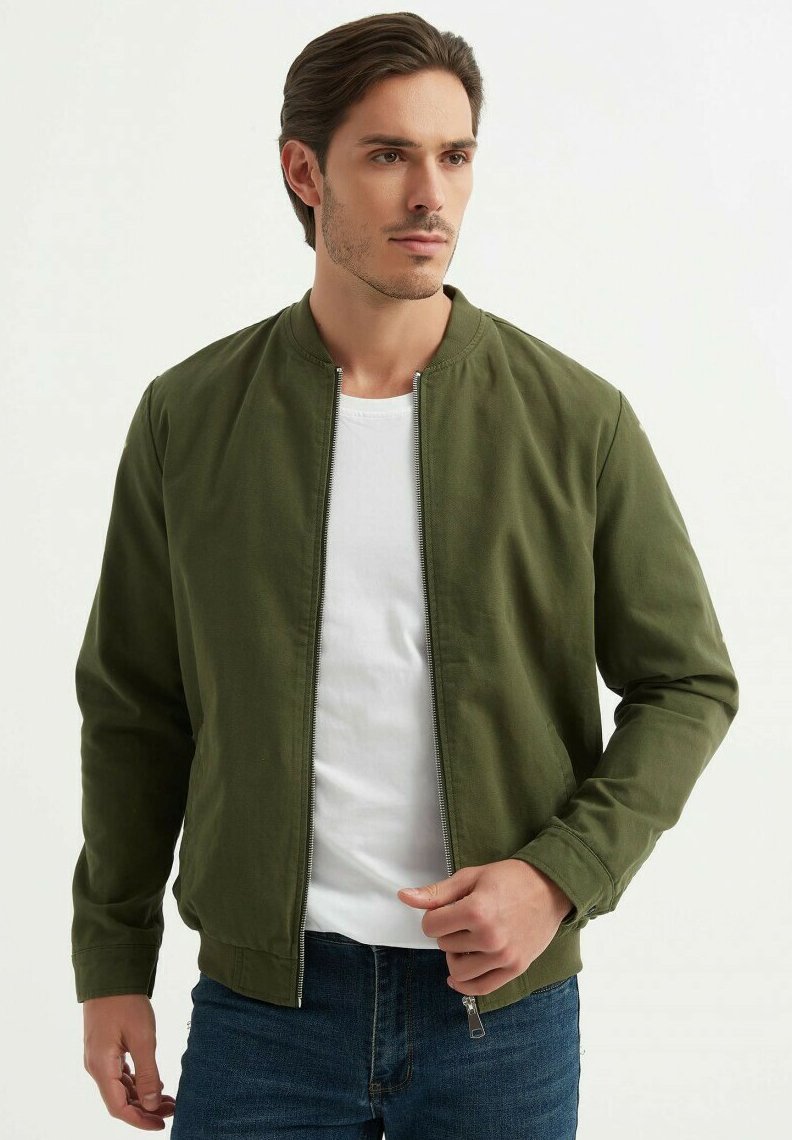 Veste bomber verte olive avec un col rond et fermeture éclair, associée à un t-shirt blanc et un jean bleu. Tissu en coton doux, design minimaliste.
