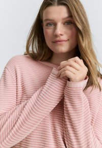 Rosa Strickpullover mit horizontalen Streifen und gerippten Bündchen, mit einem lockeren Ausschnitt und weichem Textil. Modell posiert mit gefalteten Händen.