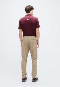 Μπορντό πόλο μπλούζα με λογότυπο στην πλάτη, σε συνδυασμό με μπεζ παντελόνι cargo με πλαϊνές τσέπες και ελαστική μέση, φορεμένο με μαύρα παπούτσια.