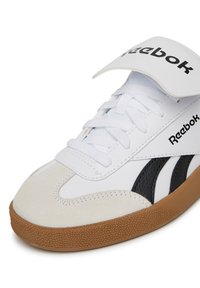 Biele športové topánky s semišovými a koženými akcentmi, s čiernym logom a pruhmi, plochými šnúrkami a štruktúrovanou gumovou podrážkou.