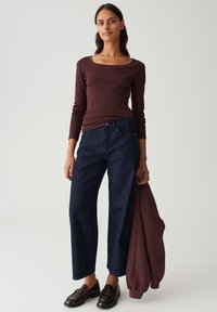 Woman wearing a burgundy long-sleeve top, dark blue wide-leg jeans, dark brown loafers, holding a matching burgundy jacket.