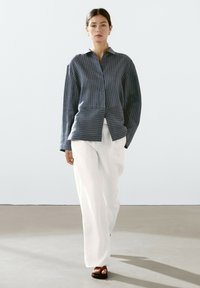 Navy striped button-up shirt with long sleeves, paired with white pinstriped wide-leg pants and brown heeled sandals on a neutral floor.