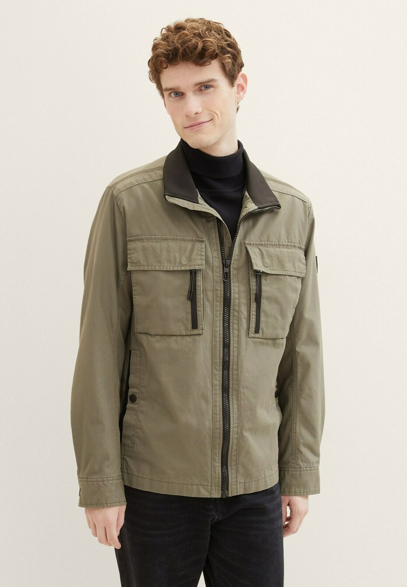 Chaqueta verde oliva con un cuello negro, cremallera frontal y múltiples bolsillos. Lleva puesta sobre un cuello de tortuga negro y combinada con jeans oscuros.