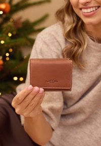 Moteris laiko rudą odinį piniginę su "Gusti Leder studio" logotipu, vilkėdama smėlio spalvos megztinį, o fone yra išblurintos Kalėdų eglutės lemputės.