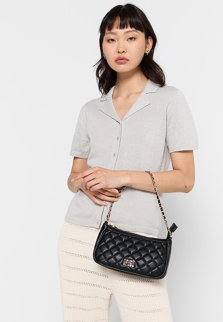 Borsa a tracolla trapuntata nera con catena, caratterizzata da una forma rettangolare e un accento con logo dorato. Il modello indossa una camicia grigia chiaro con bottoni.