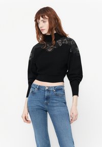 Femme aux cheveux roux portant un pull noir court avec des détails en dentelle sur les épaules et un col montant, associé à un jean bleu.
