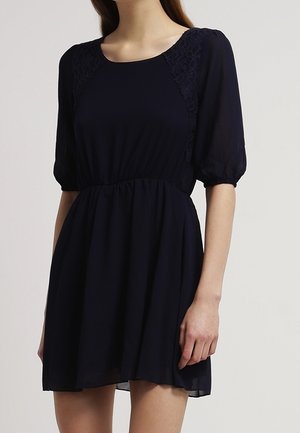 Femme portant une courte robe noire avec des manches mi-longues bouffantes et des détails en dentelle sur les épaules, debout devant un fond uni.