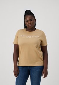 Béžové bavlnené tričko s krátkymi rukávmi a okrúhlym výstrihom, s bielym logom "TOMMY HILFIGER" a "EST. 1985." Pohľad z prednej strany.