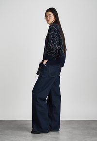 Pull en tricot marine avec des motifs géométriques, associé à un jean en denim foncé à jambes larges. Le modèle porte des lunettes à monture transparente et tient un sac noir.