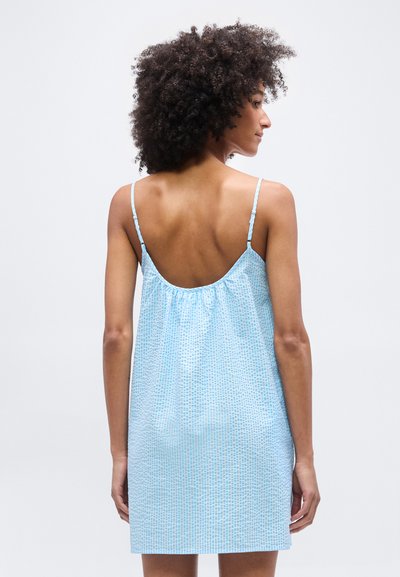 Γυναίκα με μπούκλες, που φοράει ανοιχτό μπλε αμάνικο φόρεμα με λεπτές τιράντες, γυρισμένη με την πλάτη, μπροστά σε απλό γκρι φόντο.