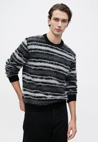 Young man with dark hair wearing a black and gray fuzzy striped sweater and black pants against a plain light background.