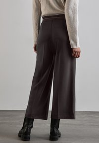 Pantaloni a gamba larga in tessuto grigio scuro, con una vita testurizzata e dettagli intricati. Indossati con stivaletti neri.