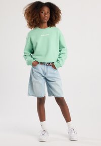 Felpa verde menta con testo bianco, abbinata a pantaloni corti di jeans azzurri. Modello che indossa sneakers bianche e calze alla caviglia.