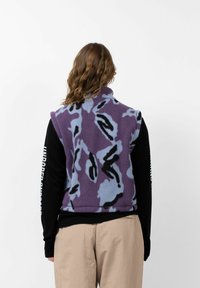 Paarse fleece vest met een blauw en zwart abstract patroon, hoge kraag, ritssluiting en zijzakken, gedragen over een zwart shirt met lange mouwen.