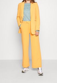 Žluté sako na míru a kalhoty s širokými nohavicemi z hladké látky, doplněné světle modrým žebrovaným svetrem a sportovními teniskami.