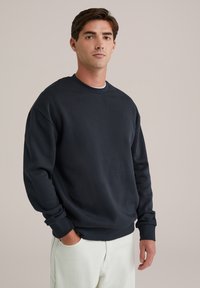 Granatowy sweter z miękkiego materiału, z klasycznym okrągłym dekoltem i prążkowanymi mankietami; noszony z jasnymi spodniami.