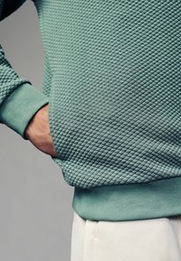 Sudadera de color verde menta con textura, patrón de diamante, puños acanalados y un corte relajado, que incluye un bolsillo lateral.