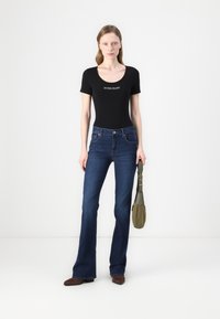 Body aderente nero con logo "GUESS JEANS", abbinato a jeans a zampa d'elefante blu scuro e stivaletti marroni a punta. La modella tiene una borsa verde.