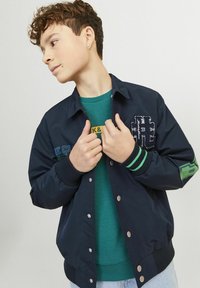 Giacca bomber blu navy con dettagli verde e grigio, dotata di logo ricamato e bottoni a pressione, indossata sopra un maglione verde e jeans chiari.