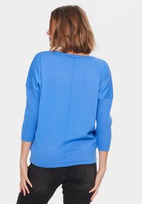 Blå tröja med trekvartsärmar, en avslappnad passform, ribbad textur på ärmarna och en slät kropp. Sömmarna går längs ryggen.