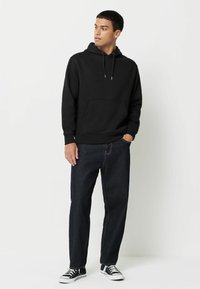 Sort bomulds hættejakke med en frontlomme, parret med mørkeblå jeans. Model iført sort og hvide sneakers. Afslappet fit design.