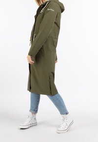 Parka longue vert olive avec capuche, fermetures éclair et cordons de serrage, comprenant des fentes latérales et deux poches avant, associée à un jean bleu et des baskets blanches.