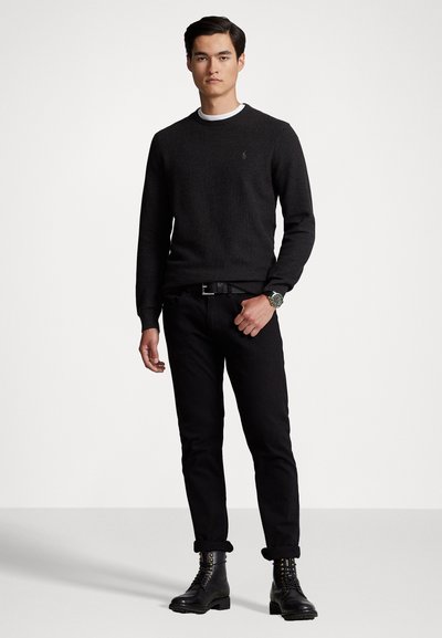 Νέος άνδρας που στέκεται φορώντας μαύρο πουλόβερ, μαύρο παντελόνι τα ψαλίδια στο ύψος του αστραγάλου, μαύρες δερμάτινες μπότες και ένα ρολόι στον καρπό, μπροστά σε απλό λευκό φόντο.