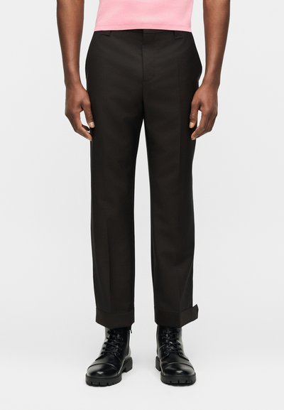 Άνδρας που φοράει μαύρο παντελόνι με ίσια μπατζάκια, γυρισμένο στον αστράγαλο, συνδυασμένο με μαύρα μποτάκια με κορδόνια και ροζ πουκάμισο.