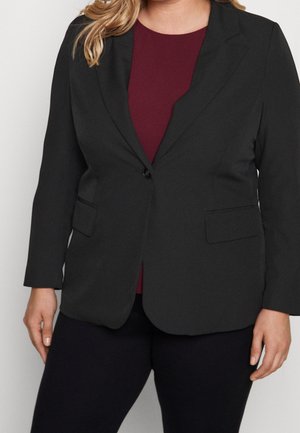 Žena v černém jednospuvodovém blejzru přes vínový top a černé kalhoty, stojící s uvolněnýma rukama podél těla.