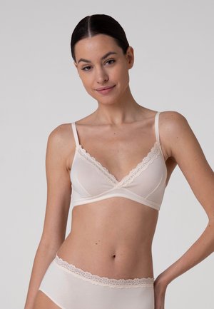Donna con capelli scuri indossa un reggiseno rosa chiaro con bordatura in pizzo e biancheria intima coordinata, in piedi su uno sfondo grigio uniforme.