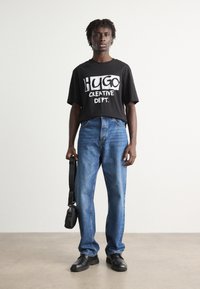 Black cropped t-shirt with "HUGO CREATIVE DEPT." in white print, blue denim jeans, black shoes, and a small black bag. Smooth textures, relaxed fit.