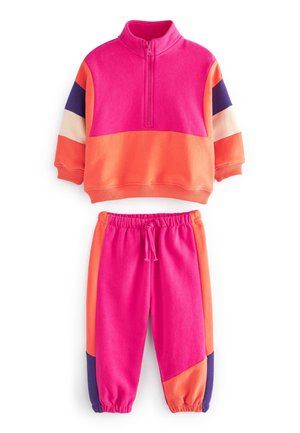 Barn iført farverigt træningssæt i pink, orange og lilla samt hvide høje sneakers går ved blomstrende buske under en klar blå himmel.