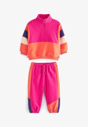 Barn iført farverigt træningssæt i pink, orange og lilla samt hvide høje sneakers går ved blomstrende buske under en klar blå himmel.
