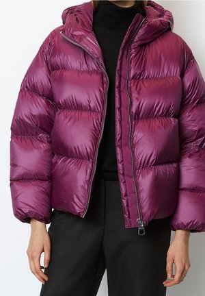 Frau trägt eine glänzende lila Steppjacke mit Kapuze, teilweise geöffnet, über einem schwarzen Rollkragenpullover und schwarzer Hose.