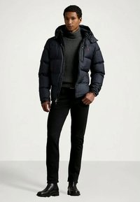 Νεαρός άνδρας που φοράει μια μαύρη μπουφάν με κουκούλα, γκρίζο πουλόβερ με ζιβάγκο, μαύρο παντελόνι και μαύρες μπότες, στέκεται μπροστά σε έναν απλό φόντο.