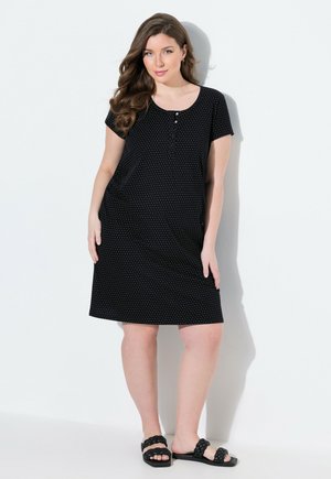 Vrouw met lang bruin haar, gekleed in een zwarte jurk met korte mouwen tot op de knie, bedrukt met witte stippen, en zwarte gevlochten sandalen, staande tegen een witte achtergrond.