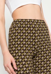 Leggings castanhos com padrão, apresentando detalhes em verde e creme. O tecido é macio e elástico, com um ajuste justo e um cós de altura média.