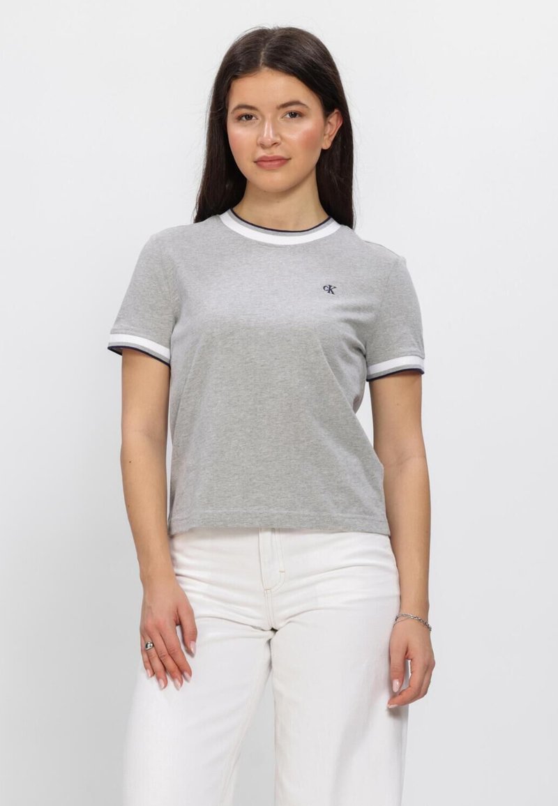 Femme aux cheveux longs et foncés portant un t-shirt gris avec des bordures blanches et noires, et un pantalon blanc, se tenant devant un fond uni.