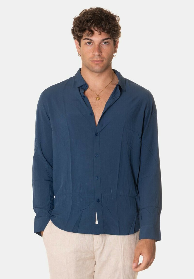 Camicia blu navy con bottoni in un tessuto leggero, caratterizzata da maniche lunghe, colletto e dettagli con taschini frontali, abbinata a pantaloni chiari.