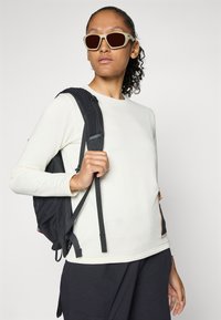 Camisa de manga larga de color crema con textura de canalé, combinada con una mochila negra. Gafas de sol con montura beige. El fondo es blanco liso.