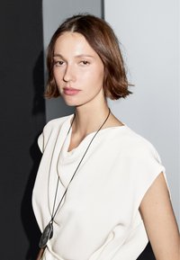 Hvid draperet top med korte ærmer, prydet med en sort pendantsmykke. Minimalistisk design, glat tekstur, blødt stof. Neutral baggrund.