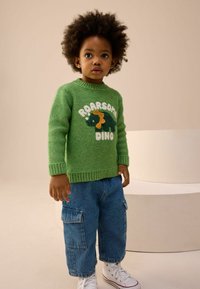 Zelený pletený sveter s textom "Roarsome Dino" a grafikou dinosaura. Kombinovaný s modrými džínovými nohavicami so zásuvkami a bielymi teniskami. Štruktúrované pletenie.