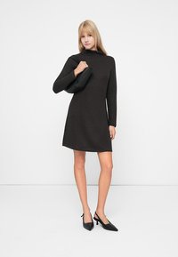 Γυναίκα με μαύρο φόρεμα μακρυμάνικο κρατώντας μια μαύρη τσάντα χεριού στον ώμο, φορώντας μαύρα ψηλοτάκουνα παπούτσια slingback με μυτερή μύτη, στέκεται μπροστά σε έναν λευκό τοίχο.