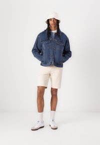 Moška jeans jakna temno modre barve, ki ima dve žepni na prsih, zapiranje na gumbe in sproščen kroj. Uparjena s smetanastimi kratkimi hlačami in belimi športnimi copati.