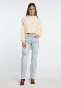 Geknipte, geribde crèmekleurige trui met hoge hals; lichtblauwe versleten jeans met rafels aan de zoom; bruine enkellaarsjes; model staat tegen een effen achtergrond.