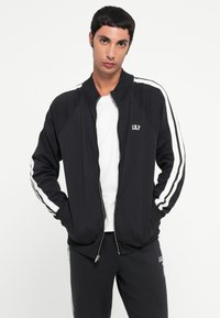 Giacca nera con zip e strisce laterali bianche, realizzata in materiale morbido. Presenta un colletto alto e il logo ricamato "GAP" sul petto.